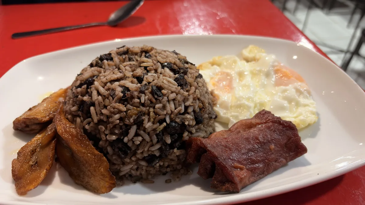 Desayuno con gallo pinto, huevo frito, plátano maduro frito y salchichón, en un plato blanco